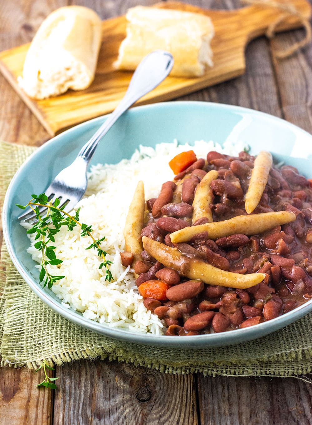 Jamaican stew peas with spinners, carrots, on steamed rice in a blue bowl, stainless steel fork, garnished with thyme sprigs on a brown wooden background with cutting board and bread