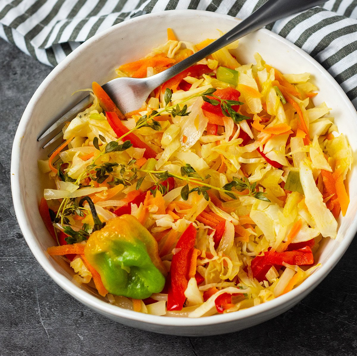 Overlay steamed cabbage, carrots, bell pepper, garnished with pepper, thyme. On a grey background with black and white stripe napkin and silver fork