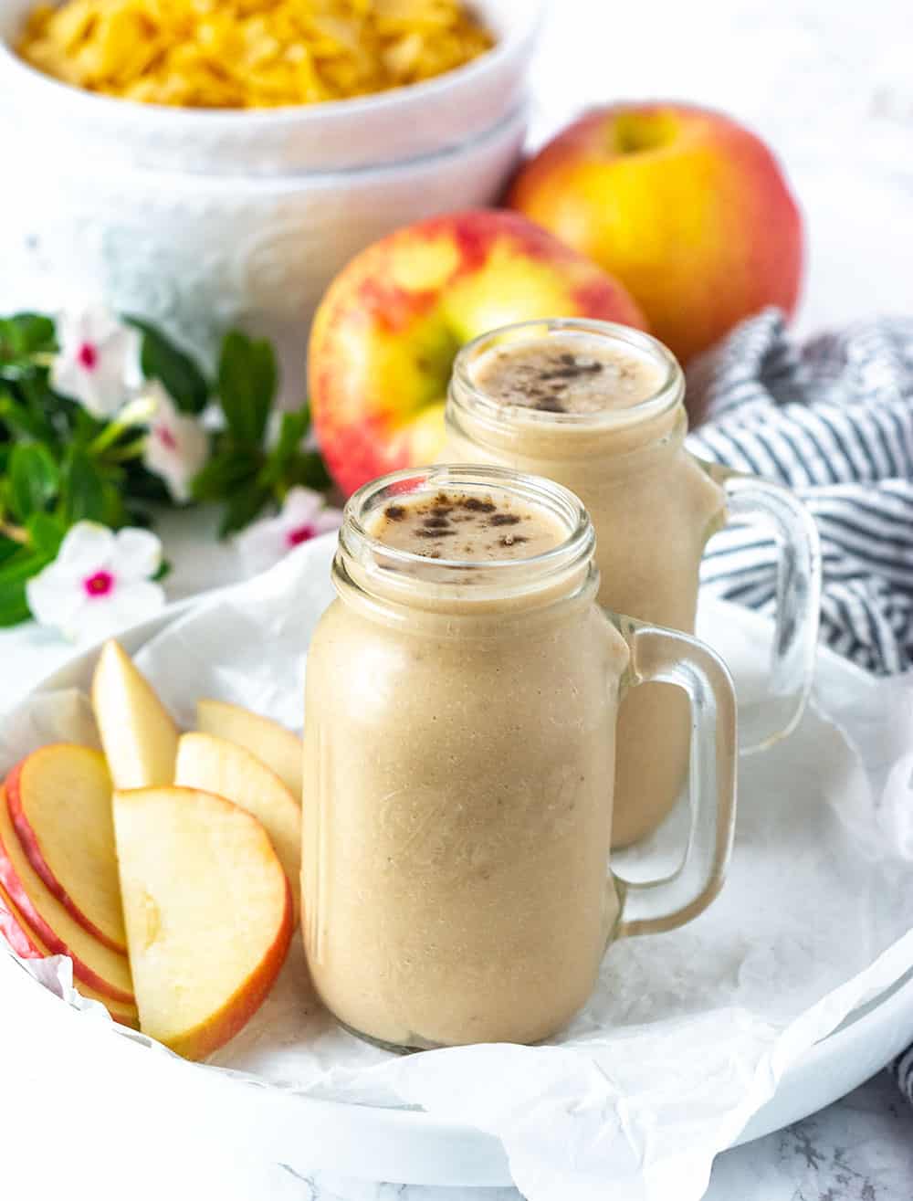 Easy Apple Pie Smoothie in a glass jar with apple slices on a white plate. apples in the background with a white bowl of cereal