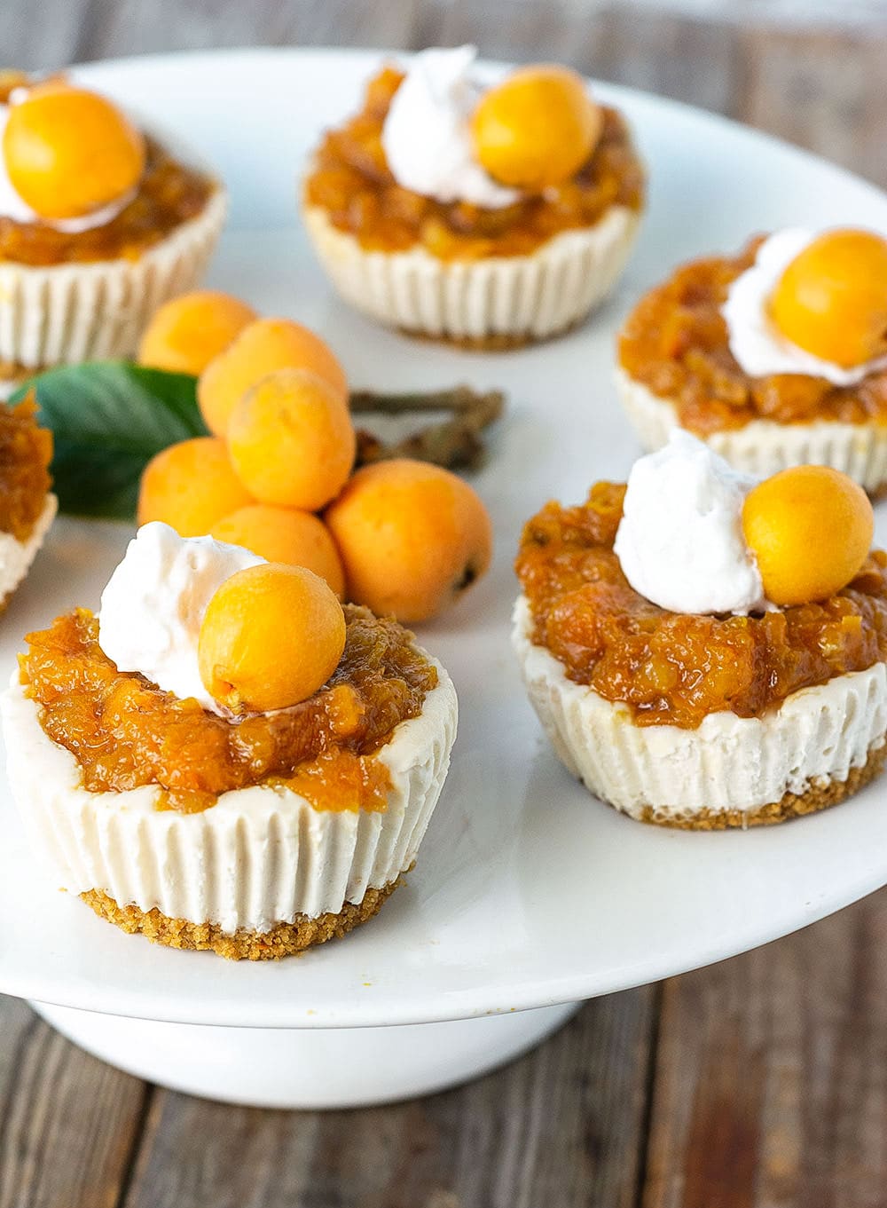 Loquat vegan cheesecake, creamy white filling on a graham cracker crust, topped with orange loquat jam, white whipped cream and fresh loquat on a white cake stand on a wooden background surrounded by fresh loquats and a green loquat leaf