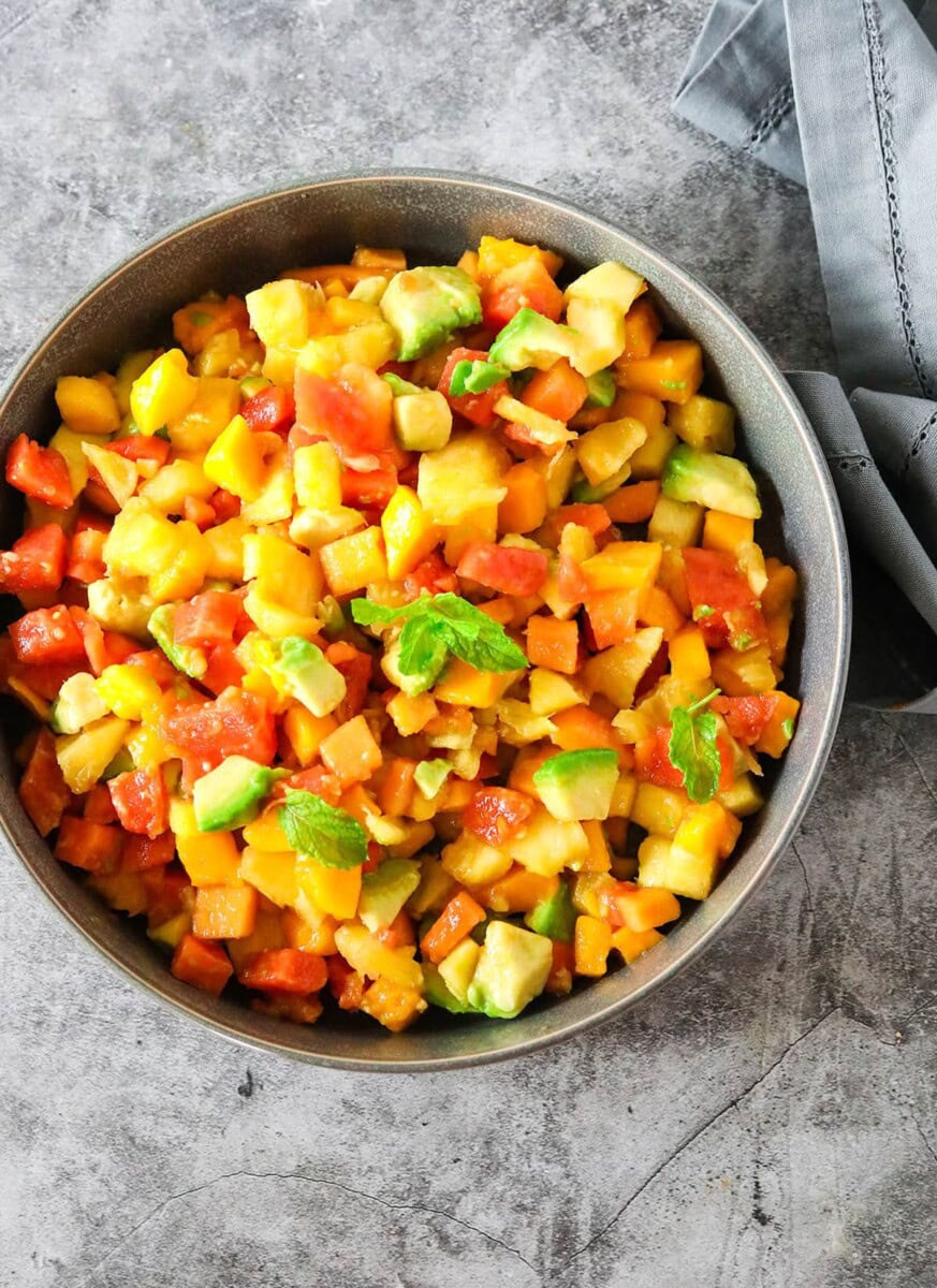 overhead papaya salad with mango, avocado, pineapple garnished with mint on a grey background