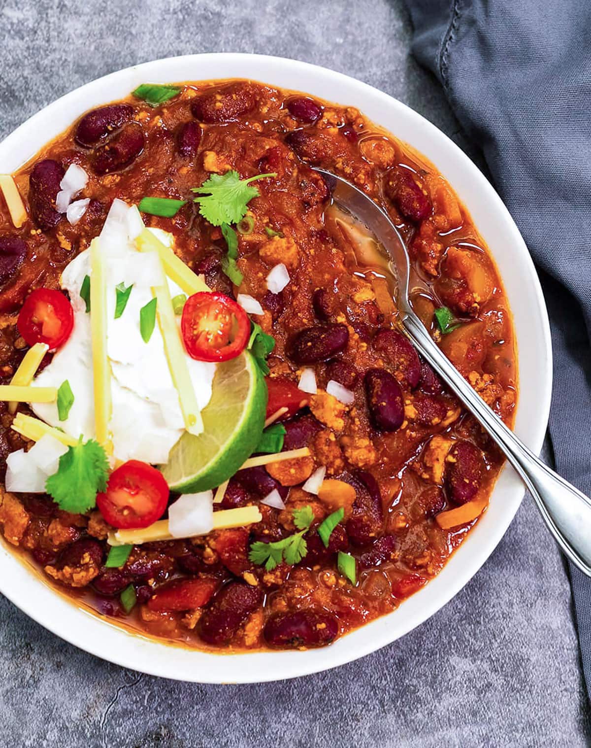 Overlay vegan chili in a white bowl garnished with a dollop of vegan sour cream, cheese shreds, tomato, green onion, cilantro, lime slice
