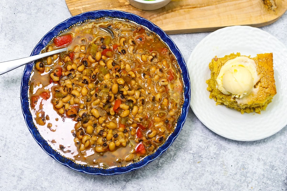 black eyed peas in a blue bowl next to a white plate with a slice of cornbread on white concrete background